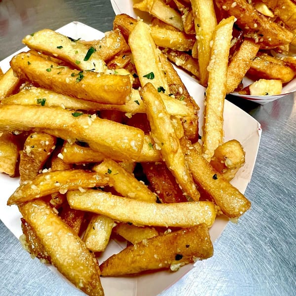 Two servings of golden-brown Frymazing Korean fries, topped with garlic and green herbs, in paper trays.