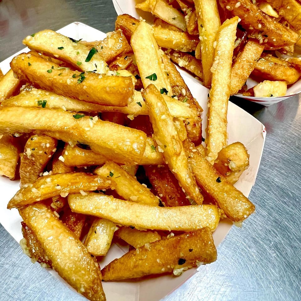 Two servings of golden-brown Frymazing Korean fries, topped with garlic and green herbs, in paper trays.